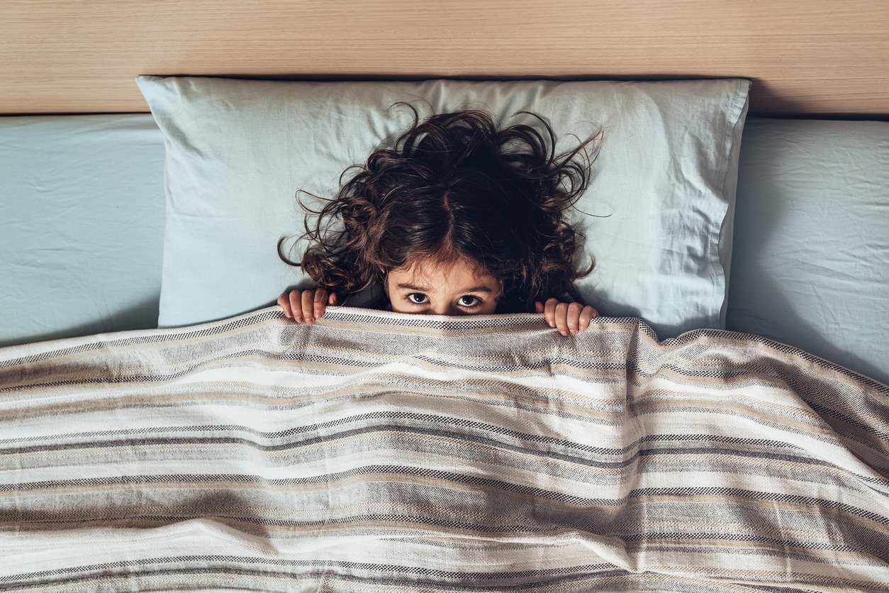 little girl covering her face with a bed sheet, only lets you see her eyes looking at the camera, happy childhood and healthy rest concept
